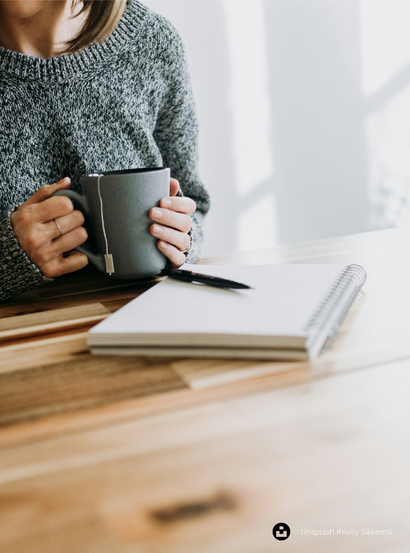 A person in a cozy knit sweater holding a warm mug, with a notebook and pen nearby — a quiet moment of reflection